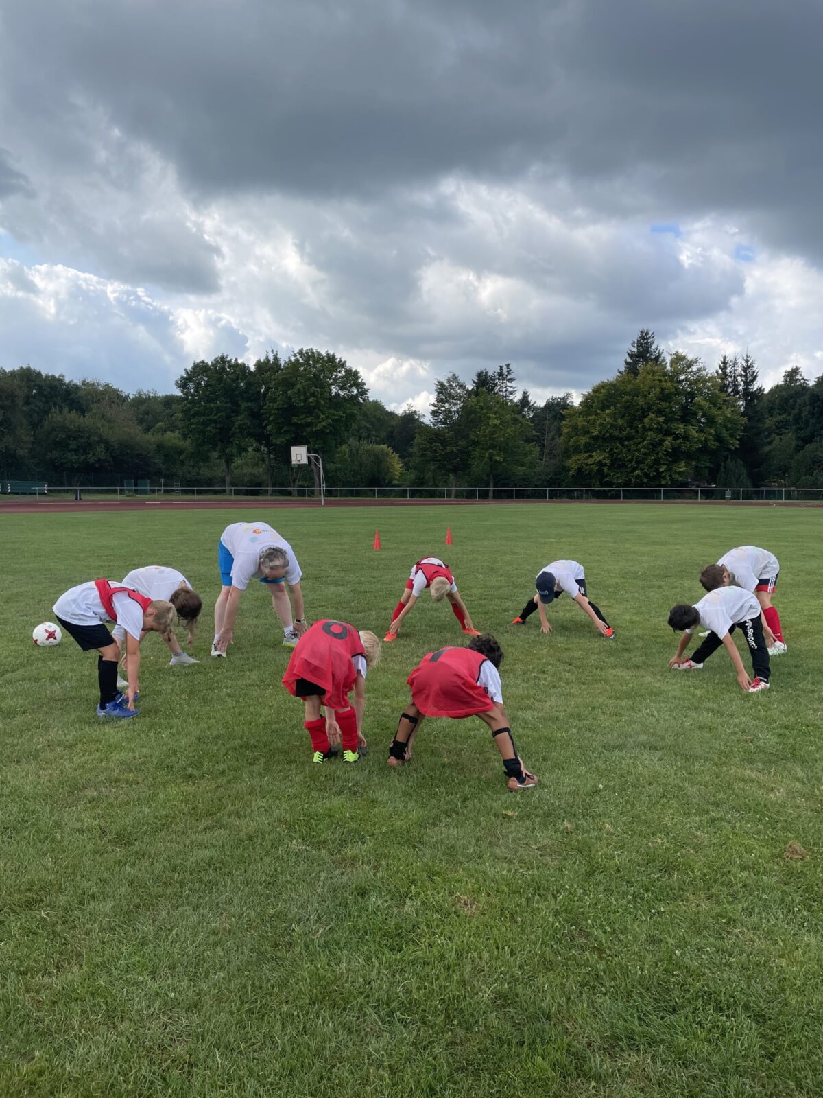 Eine Gruppe Kinder und ein Trainer stehen im Kreis auf einer Wiese. Sie stehen mit den Beinen gespreizt und dehnen ihren Oberkörper nach vorne.