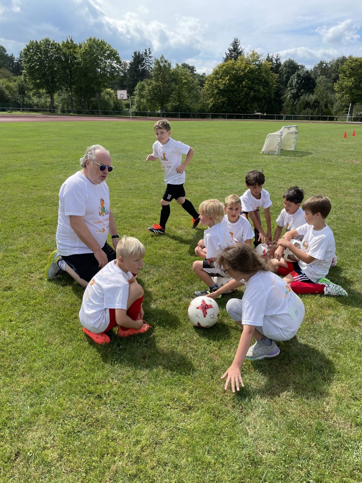 Eine Gruppe Kinder kniet auf dem Boden vor einem Mann auf einer Wiese. Sie tragen alle weiße T-Shirts mit dem KiNiKi Gecko drauf und tragen Fußballklamotten. Im Hintergrund sind Bäume und der blaue Himmel zu sehen.
