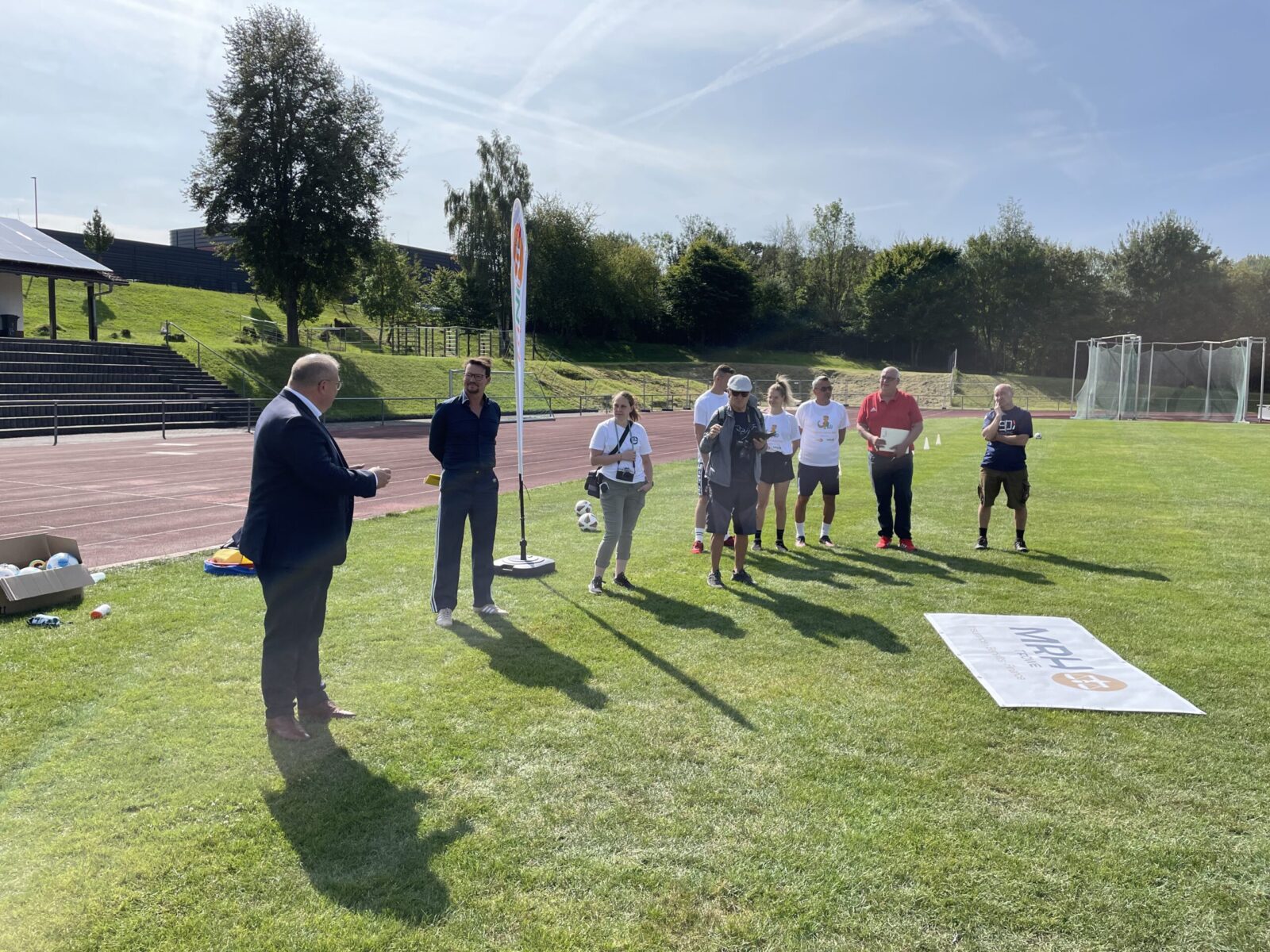 Eine Gruppe Erwachsene stehen auf einem Fußballfeld. Vor Ihnen liegt ein Banner mit dem MRH Trowe Logo drauf.
