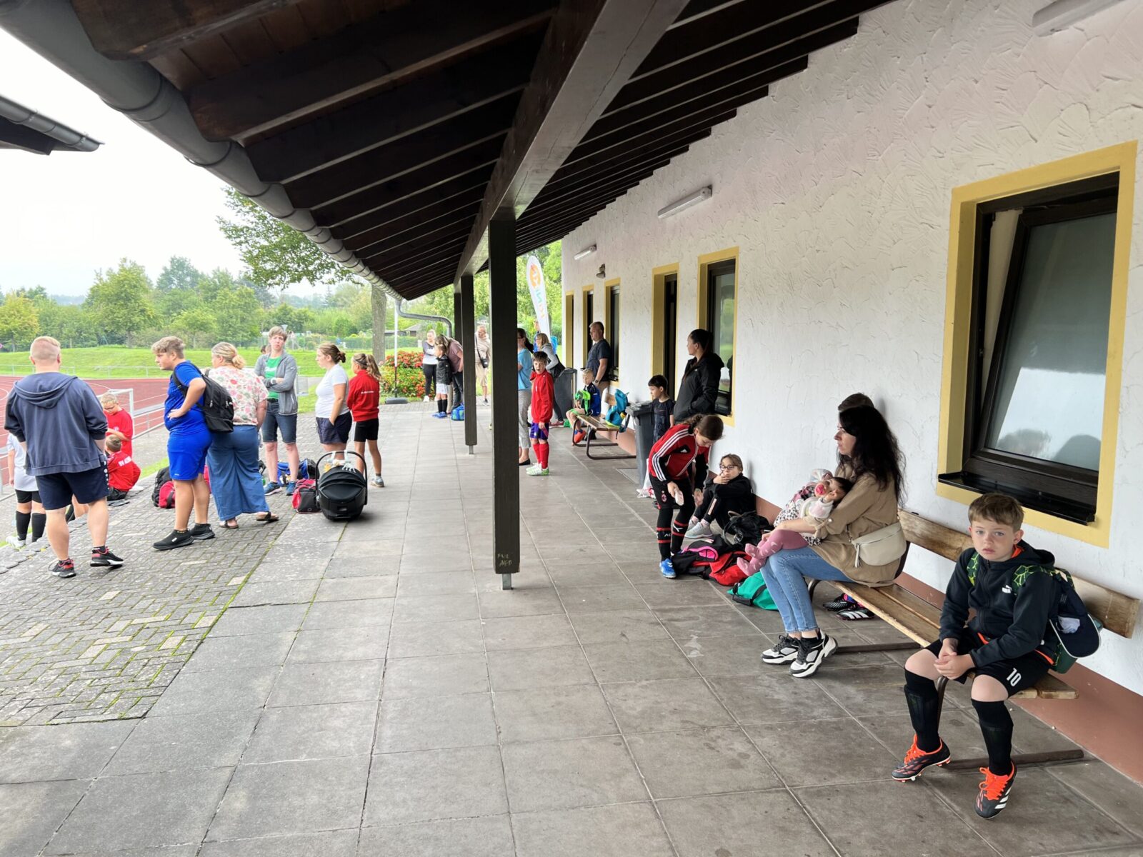 Mehrere Erwachsene und Kinder, teils in Fußballkleidung, warten und unterhalten sich vor einem langgestreckten weißen Gebäude mit überdachtem Eingangsbereich. Einige sitzen auf einer Bank, andere stehen. Im Hintergrund ist ein Sportplatz zu sehen.