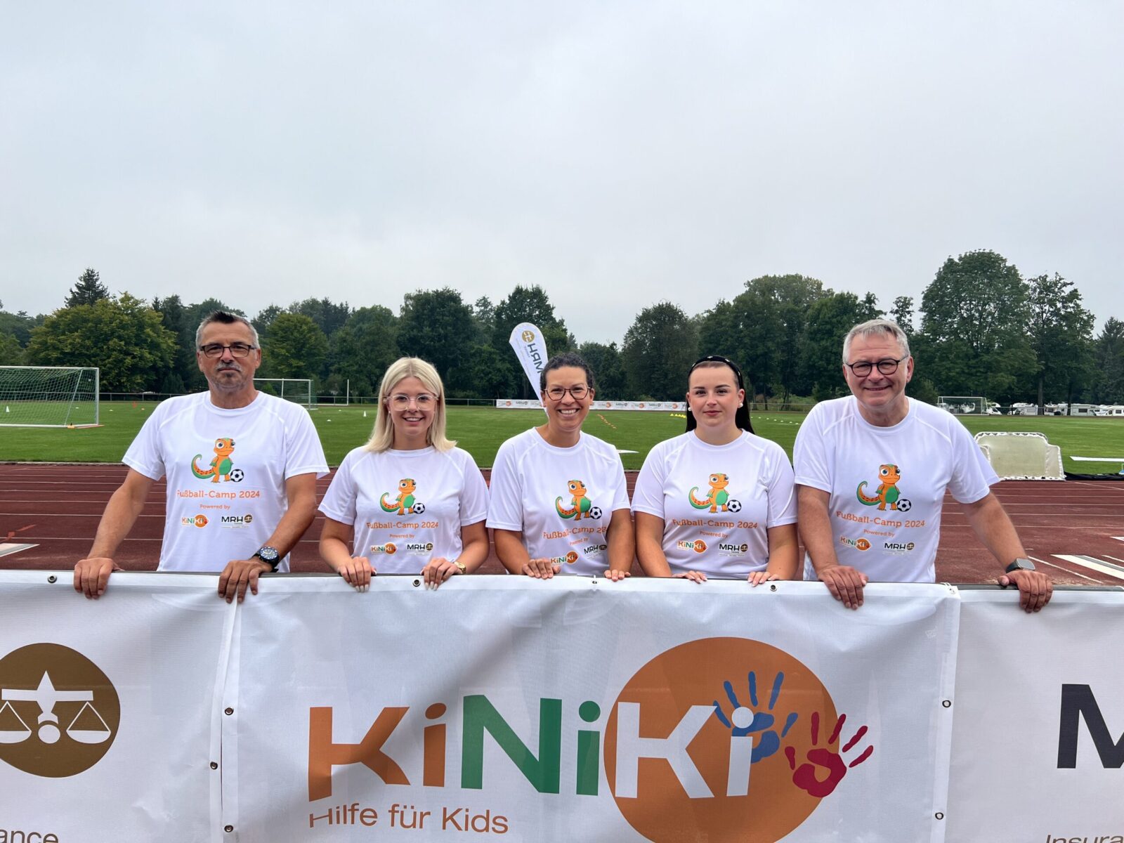 Erwachsene stehen in weißen T-Shorts mit dem KiNiKi Gecko drauf vor einem Zaun, an dem Banner mit dem KiNiKi und MRH Trowe Logo drauf hängen. Hinter Ihnen ist ein Fußballfeld zu sehen.