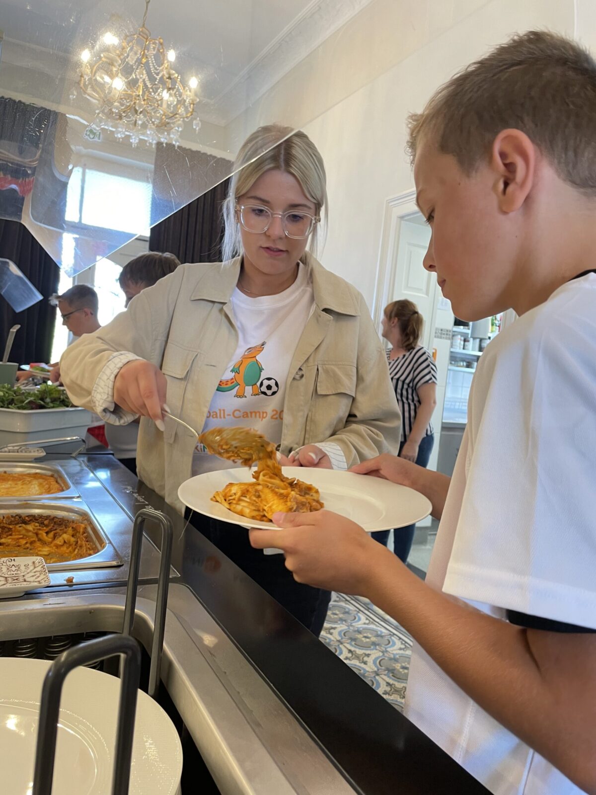 In einer Kantine oder einem Speisesaal steht eine junge Frau hinter einer Essensausgabe und reicht einem Jungen eine Portion warmes Essen. Der Junge hält ein Tablett in den Händen. Weitere Kinder stehen in der Schlange und warten darauf, bedient zu werden. Das Ambiente wirkt freundlich und einladend, mit großen Fenstern, durch die Tageslicht einfällt.