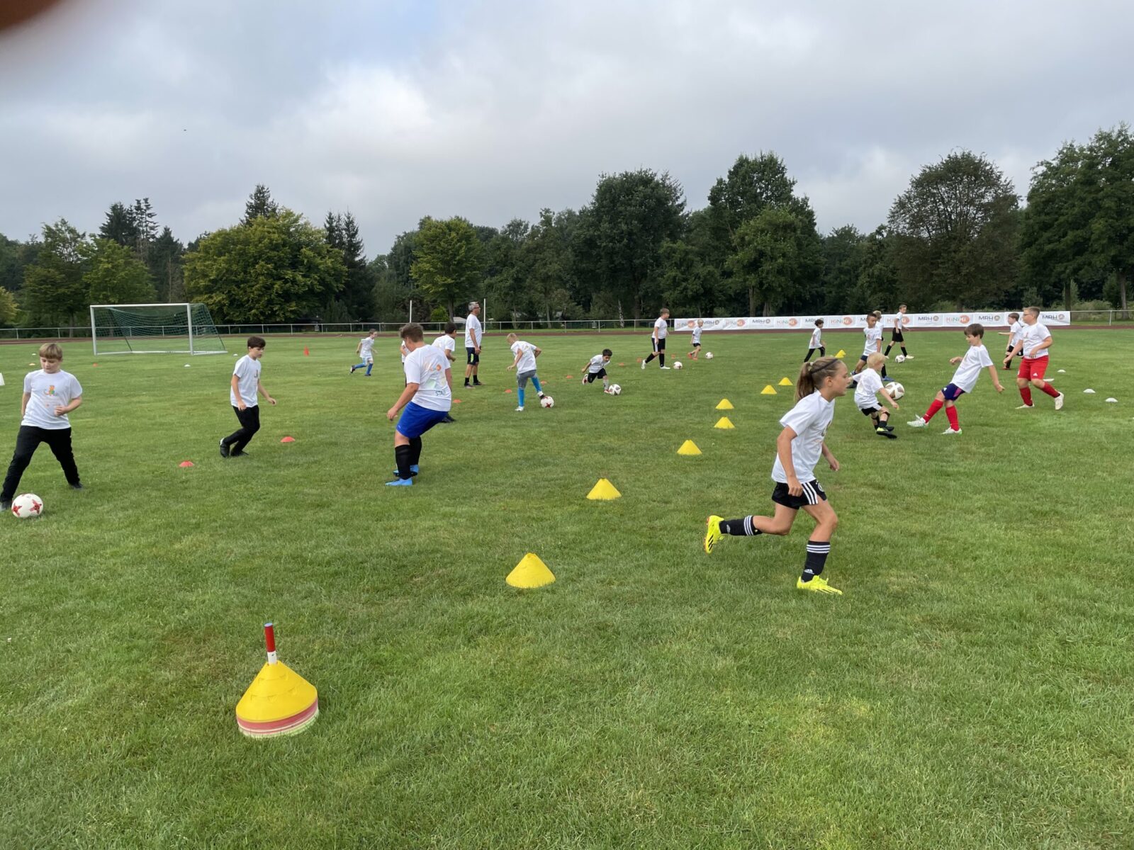 Mehrere Kinder in weißen T-Shirts und Sportkleidung rennen über eine große Wiese und spielen Fußball. Gelbe Hütchen sind auf dem Feld verteilt, um Spielfeldgrenzen oder Übungsbereiche zu markieren. Einige Kinder sprinten dem Ball hinterher. Der Himmel ist leicht bewölkt.