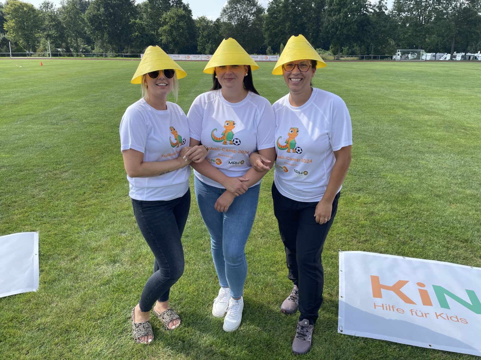 Drei Frauen stehen auf einer grünen Wiese und tragen weiße T-Shirts mit dem KiNiKi-Logo. Sie haben auffällige gelbe Hüte aufgesetzt. Sie lachen fröhlich in die Kamera, während sie entspannt nebeneinander posieren. Im Hintergrund sieht man eine große Wiese mit weiteren Veranstaltungsteilnehmern.