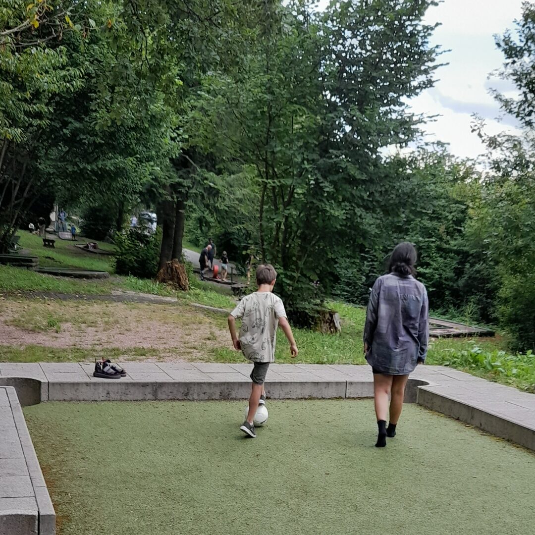 Zwei Kinder laufen in einem Park auf einem kunstrasen Feld. Das eine Kind hat einen Fußball.