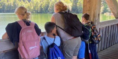 4 Kinder und 1 Frau stehen angelehnt an einem Holzrahmen auf einer Brücke und schauen auf einen See. Es ist ein sommerlicher Tag und die Sonne scheint.