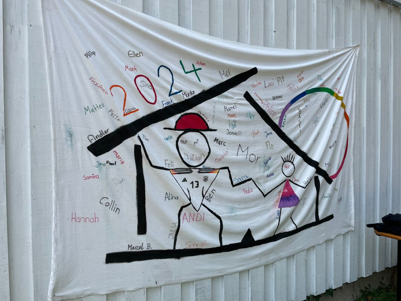 Ein weißes Lacken hängt an einem weißen Container. Auf dem Lacken sind Zeichnungen von Fußballer-Strichmännchen und darüber steht "2024". Zudem sind einige Unterschriften drauf verewigt.