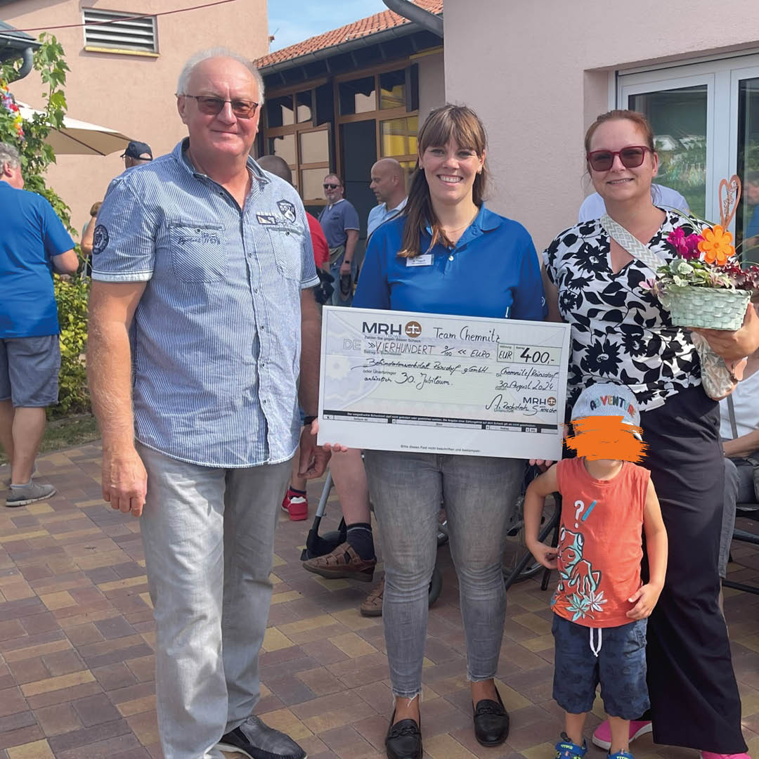 Das Bild zeigt eine kleine Gruppe von vier Personen: ein Mann, eine Frau in blauer Kleidung, eine Frau mit einer Blumenstrauß in der Hand und ein kleiner Junge mit rotem T-Shirt und Kappe. Die Frau in der Mitte hält einen symbolischen Scheck über 400 Euro, während alle in die Kamera lächeln. Es handelt sich um eine Spendenübergabe. Im Hintergrund sind weitere Personen in einer entspannten Außenumgebung zu sehen.