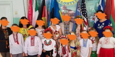 Eine Gruppe an Kindern mit 7 Erwachsenen stehen in einem Raum an einer Wand und lächeln in die Kamera. Hinter Ihnen sind verschiedene Flaggen zu sehen, unter anderem die Flagge der Ukraine und auch die Deutschland Flagge. Die Kinder scheinen typisch ukrainische Kleidung zu tragen und die Erwachsenen ebenfalls.