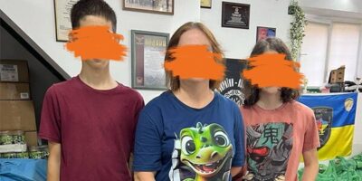 Eine Frau mit zwei Jugendlichen steht in einem Raum, der offenbar ein Lager für Hilfsgüter ist. Die Frau hält eine grüne Plastiktüte mit Hilfsgütern in der Hand. Im Hintergrund sind weitere grüne Tüten und Kartons mit Vorräten zu sehen. An der Wand hängen verschiedene Urkunden und eine ukrainische Flagge.