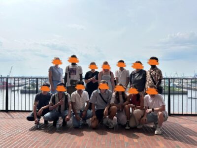 Das Bild zeigt eine Gruppe von 14 Personen, die draußen vor einer Brüstung posiert, mit einem Wasserhafen oder einer Küstenlandschaft im Hintergrund. Einige Personen sitzen auf dem Boden, während andere stehen. Die Kleidung ist sommerlich und die Atmosphäre wirkt entspannt, als ob sie sich auf einem Ausflug oder einer gemeinsamen Reise befinden. Der Himmel ist leicht bewölkt, aber das Wetter scheint angenehm.