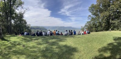 Eine Gruppe von Kindern und Jugendlichen sitzt auf einer Wiese am Waldrand und genießt den weiten Blick über ein grünes Tal und bewaldete Hügel unter einem leicht bewölkten Himmel.