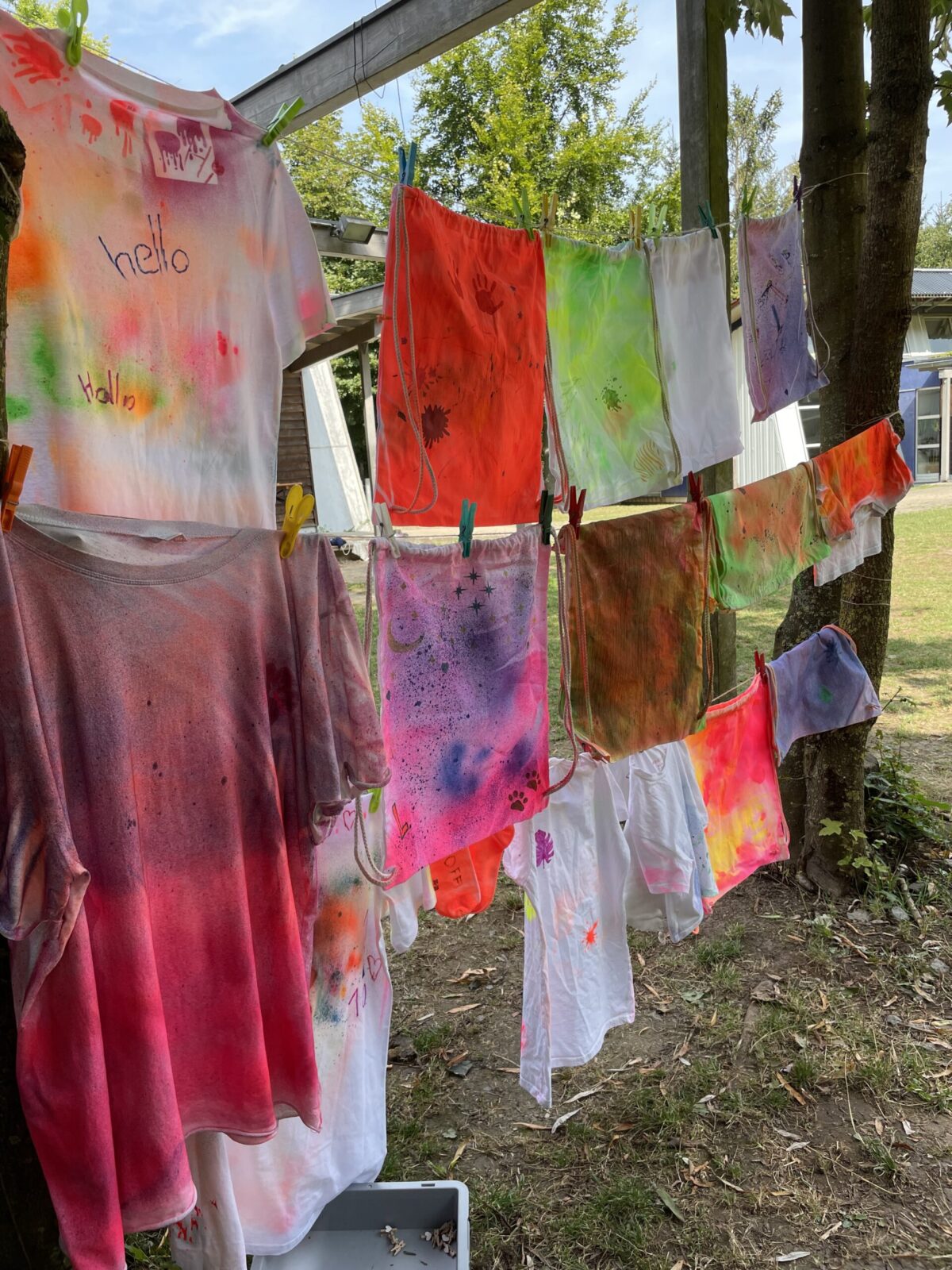 Das Bild zeigt eine Wäscheleine, an der eingefärbte T-Shirts hängen. Es ist ein sommerlicher Tag und die Sonne scheint.