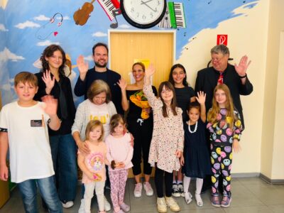 Das Bild zeigt eine Gruppe Menschen bestehend aus Kindern und Erwachsenen. Sie stehen vor einer Wand, auf der Musikinstrumente sind. Sie schauen alle lächelnd und winken mit der rechten Hand in die Kamera.