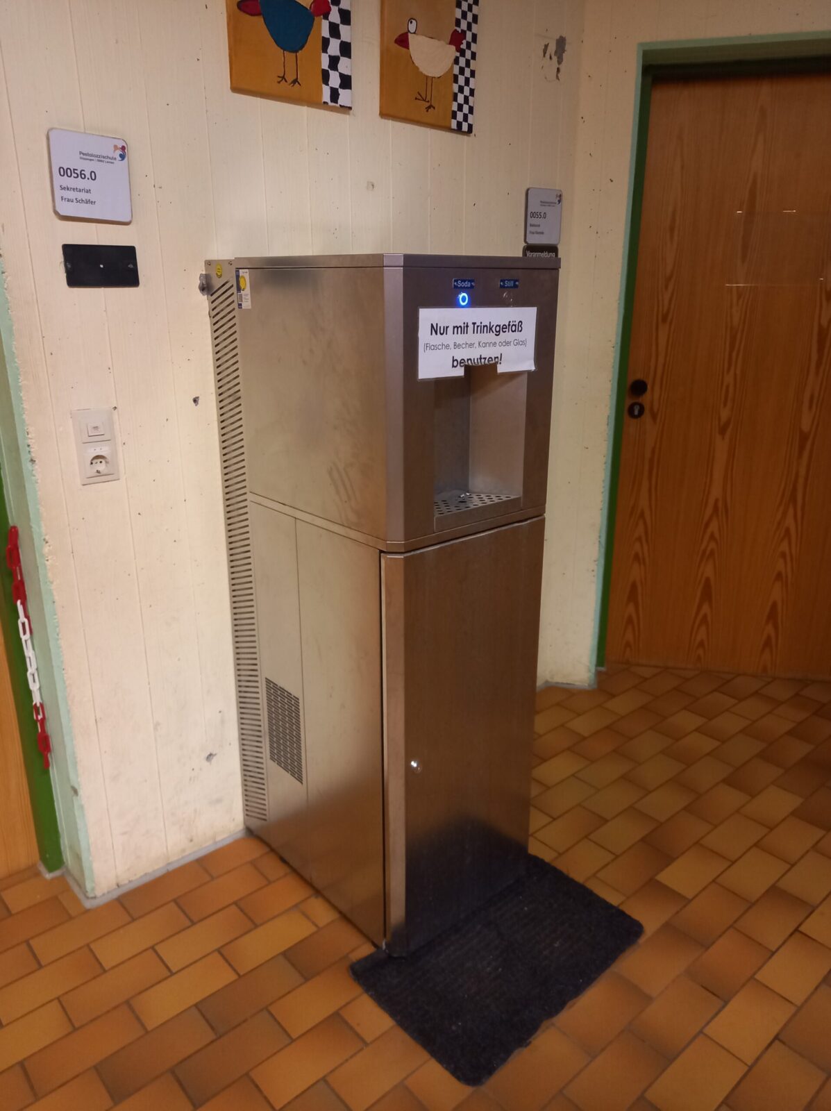 Das Bild zeigt einen Wasserspender in einem Schulflur, umgeben von heller Holzverkleidung und einer gefliesten Bodenfläche in warmen Brauntönen. An der Wand sind mehrere Schilder und bunte Kinderzeichnungen angebracht. Auf dem Wasserspender befindet sich ein Schild mit der Aufschrift „Nur mit Trinkgefäß (Flasche, Becher, Kanne oder Glas) benutzen“. Der Wasserspender wirkt robust, aber schon etwas abgenutzt, was darauf hindeutet, dass er schon länger im Einsatz ist.