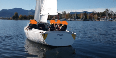Ein Segelboot mit Schülern, das auf einem ruhigen See fährt. Im Hintergrund sind die Chiemgauer Alpen und der Hafen von Prien zu sehen.