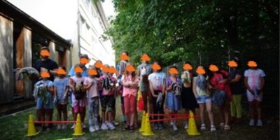 Eine Gruppe von Kindern steht mit Steckenpferden in einem schattigen Außenbereich. Sie sind vor einer Reihe von kleinen Hindernissen aufgestellt, die aus Hütchen und Stangen bestehen. Die Kinder tragen fröhliche, sommerliche Kleidung und stehen unter Bäumen, die angenehmen Schatten spenden. Im Hintergrund ist ein Gebäude zu sehen.
