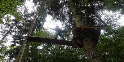 Eine Person steht auf einem schmalen Holzsteg hoch oben in einem Hochseilgarten, gesichert mit einem Klettergurt und Halteseilen. Die Person scheint konzentriert, während sie das Gleichgewicht hält, um von einer Plattform zur nächsten zu gelangen. Umgeben von hohen, dicht belaubten Bäumen, bietet die Szene einen spannenden Blick auf die Herausforderungen eines Kletterparks inmitten der Natur.