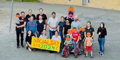 Ein Gruppenfoto mit allen Helfenden und den Kindern. Sie stehen nah beieinander und lächeln in die Kamera. In der vorderen Reihe wird das bemalte Plakat gehalten. Im Hintergrund sieht man recht die Schaukel und den Sandkasten. Links sieht man die frisch gestrichenen Beete.