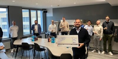 Eine Gruppe von Menschen steht in einem modernen Büro, lächelnd und mit Getränken in der Hand. Im Vordergrund hält ein Mann stolz einen symbolischen Scheck über 700 Euro, der für wohltätige Zwecke gesammelt wurde.