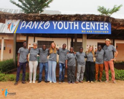 Eine Gruppe von Menschen steht vor dem PANKO Youth Center, unter einem Schild mit der Aufschrift „PANKO Youth Center - We Are Together“. Die Gruppe trägt graue T-Shirts mit dem PANKO-Logo und lächelt in die Kamera. Im Hintergrund ist das Zentrum mit einem Strohdach und einer gepflegten Umgebung zu sehen.