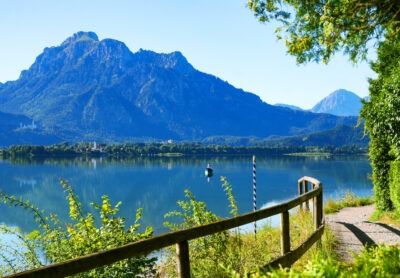 Ein ruhiger See im Ostallgäu spiegelt die beeindruckenden Berge im Hintergrund wider. Am klaren blauen Himmel scheint die Sonne, während ein schmaler Wanderweg mit einem Holzgeländer am rechten Bildrand entlangführt. Üppiges Grün und Büsche säumen den Weg, und in der Ferne ist ein kleines Boot auf dem See zu erkennen. Ein malerisches Naturpanorama, das Ruhe und Schönheit ausstrahlt.