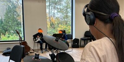 Ein Mädchen spielt konzentriert auf einem elektronischen Schlagzeug in einem modernen, lichtdurchfluteten Raum. Im Hintergrund sind zwei weitere Kinder mit Musikinstrumenten zu sehen, darunter eine Gitarre, während sie gemeinsam musizieren.