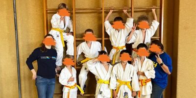 Das Bild zeigt insgesamt 8 Jungs und zwei Frauen. Sie stehen als Gruppe vor einer Holz-Kletterwand. 4 Jungs hängen an an der Kletterwand. Sie lächeln alle freundlich in die Kamera und manche zeigen mit ihren Fingern "Peace".