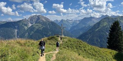 Ein schmaler Wanderpfad führt zwei Personen entlang einer grünen Bergwiese hin zu einem Gipfelkreuz. Im Hintergrund erstreckt sich eine beeindruckende Alpenlandschaft mit schroffen Berggipfeln und einem strahlend blauen Himmel mit weißen Wolken.