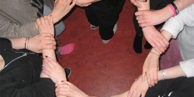 Eine Gruppe von Personen bildet einen Kreis, indem sie sich gegenseitig an den Unterarmen festhalten. Die Szene symbolisiert Gemeinschaft, Zusammenhalt und gegenseitige Unterstützung. Die Hände und Arme der Personen stehen im Mittelpunkt, während der Hintergrund in warmen Rottönen gehalten ist.