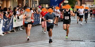 Eine Gruppe von Läufern, darunter ein junger Teilnehmer, läuft während eines Stadtlaufs durch die Innenstadt. Die Zuschauer am Rand feuern die Läufer an, während die Teilnehmenden unter einem Start-/Zielbogen hindurchlaufen. Die Atmosphäre ist lebendig, mit Jubel und sportlichem Einsatz.