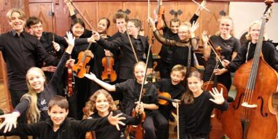 Eine Gruppe von Jugendlichen mit Musikinstrumenten wie Geigen, Cello und Kontrabass posiert fröhlich in einem Konzertsaal. Alle tragen schwarze Kleidung und strahlen Freude und Begeisterung aus, während sie ihre Instrumente hochhalten. Im Hintergrund sind Holzverkleidungen und eine warme, einladende Atmosphäre zu sehen.