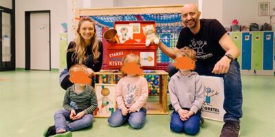 Eine fröhliche Szene in einer Kita: Zwei Erwachsene – eine Frau und ein Mann – sitzen zusammen mit drei Kindern vor der „Starken Kinder Kiste!“. Die rote Präventionskiste ist mit Materialien gefüllt und mit den Logos der Deutschen Kinderschutzstiftung Hänsel+Gretel und KiNiKi versehen. Die Kinder sitzen auf kleinen Hockern, während im Hintergrund ein heller, kindgerechter Raum mit Spinden und Spielgeräten zu sehen ist. Der Mann hält ein Kuscheltier hoch, und alle lächeln fröhlich in die Kamera.