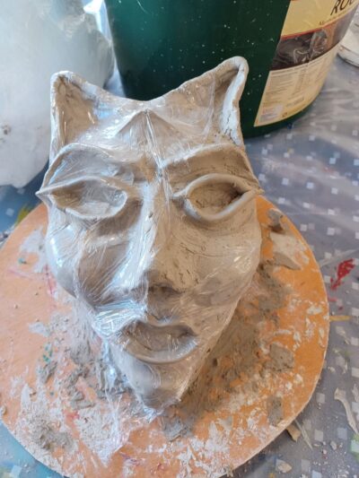 Zu sehen ist eine Maske, die aus Ton angefertigt wurde und für das Theaterstück verwendet werden soll. Die Maske hat spitze Ohren, große Augen und einen offenen Mund. Sie liegt auf einer Tonplatte auf einer Malunterlage.