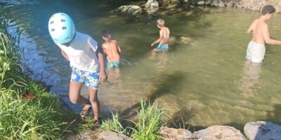 Mehrere Kinder spielen und baden in einem flachen Fluss, umgeben von grüner Natur. Eines der Kinder trägt einen blauen Helm und steigt gerade aus dem Wasser auf eine steinige Böschung. Die Sonne scheint, und das Wasser reflektiert das Licht – ein unbeschwerter Moment in der Natur.