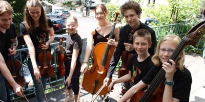Eine Gruppe Kinder steht draußen auf einem Balkon in einem Kreis. In der einen Hand halten sie jeweils eine Geige und in der anderen einen Geigenbogen. Sie tragen alle Schwarze Klamotten und lächeln freundlich in die Kamera. Im Hintergrund sind Häuser und Bäume zu sehen.