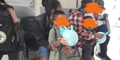 Personen - darunter 4 Erwachsene und 2 kleinere Kinder - sitzen auf schwarzen Stühlen in einem Wartezimmer.