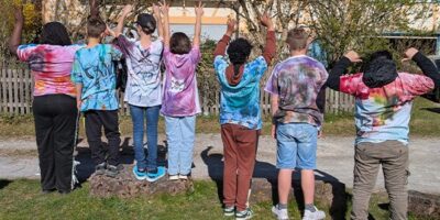 Eine Gruppe von sieben Kindern steht mit dem Rücken zur Kamera und hebt die Arme. Sie tragen bunte, selbstgestaltete T-Shirts und stehen im Freien auf Steinen, vor einem Zaun und Bäumen.