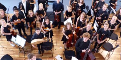 Eine Gruppe von Musikern in einem Orchester bereitet sich auf ein Konzert vor. Sie sind in einem Proberaum und tragen schwarze Kleidung. Verschiedene Instrumente wie Geigen, Celli, Kontrabässe und Blasinstrumente sind zu sehen.