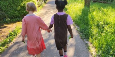 Zwei kleine Mädchen gehen Hand in Hand einen sonnigen Weg entlang, der von grünen Bäumen und Gras gesäumt ist. Beide tragen farbenfrohe Kleidung; das linke Mädchen trägt ein rosa Kleid, während das rechte Mädchen ein braunes Kleid und rosa Schuhe trägt. Die Szene strahlt Freundschaft und Unbeschwertheit aus.