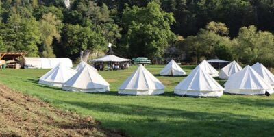 Ein Zeltlager auf einer grünen Wiese, umgeben von dichtem Wald. Mehrere weiße Zelte sind aufgestellt, während eine Gruppe von Menschen unter einem großen Pavillon versammelt ist. Im Hintergrund sind Bäume und ein Fahrzeug mit Kanus zu sehen.