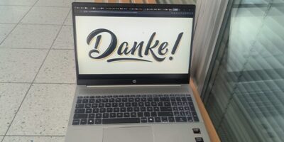 Ein silberner HP-Laptop steht auf einer Holzbank und zeigt auf seinem Bildschirm das Wort "Danke!" in stilisierter Schrift. Der Laptop befindet sich in einem modernen, hellen Raum mit Glaswänden und einem gefliesten Boden. Die Tastatur ist beleuchtet und auf dem Laptop sind zwei AMD-Aufkleber zu sehen.