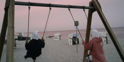 Zwei Personen schaukeln am Strand bei Sonnenuntergang. Der Himmel ist in sanften Rosa- und Lilatönen gefärbt, und im Hintergrund sind Strandkörbe sowie das Meer zu sehen. Die Szene vermittelt eine friedliche und entspannte Atmosphäre.