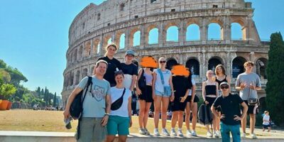 Eine Gruppe von Menschen steht vor dem Kolosseum in Rom, Italien. Das historische Amphitheater erstreckt sich majestätisch im Hintergrund. Die Personen tragen sommerliche Kleidung und stehen lächelnd nebeneinander.