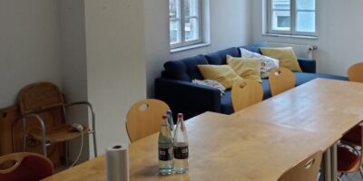 Ein Besprechungsraum mit einem langen Holztisch und mehreren Holzstühlen. Im Hintergrund befindet sich eine blaue Couch mit gelben Kissen, die vor zwei Fenstern steht. An der Decke ist ein Projektor montiert und auf dem Tisch stehen Wasserflaschen und eine Rolle Küchenpapier.