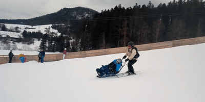 Eine Frau fährt mit Skiern einen schneebedeckten Hang hinunter und hat vor sich ein Skibob, welches Sie führt. Sie und der Junge auf dem Skibob tragen einen Helm und dicke Winterkleidung. Im Hintergrund sind weitere Personen zu sehen, die an einer Holzbarriere stehen, sowie eine bewaldete Landschaft und schneebedeckte Hügel.