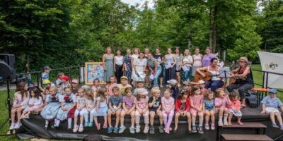 Eine große Gruppe von Kindern sitzt auf einer Bühne im Freien, während Erwachsene im Hintergrund stehen und musizieren. Die Szene spielt sich in einer grünen Parklandschaft ab, umgeben von Bäumen und sommerlichem Wetter. Die Kinder wirken fröhlich und bunt gekleidet, während die Erwachsenen eine Gitarre und Notenblätter halten, was auf eine musikalische Darbietung hinweist.