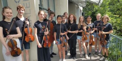 Eine Gruppe junger Musikerinnen und Musiker steht mit ihren Streichinstrumenten, darunter Geigen und Bratschen, auf einer sonnigen Terrasse. Sie tragen einheitliche schwarze T-Shirts mit einem bunten Logo und sind umgeben von grüner Natur und einem Gebäude im Hintergrund. Die Stimmung wirkt konzentriert und harmonisch, passend zu einem musikalischen Anlass.