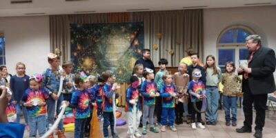 Eine Gruppe von Kindern steht in einem Raum mit dekorativem Hintergrund, der eine märchenhafte Szene mit Lichtern und Pflanzen zeigt. Die Kinder tragen bunte, tie-dye T-Shirts und halten verschiedene Requisiten in der Hand, während sie gemeinsam auf einer Veranstaltung auftreten. Im Hintergrund sind Erwachsene zu sehen, die die Situation beobachten.