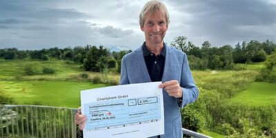 Ein Mann in einem blauen Sakko steht auf einer Terrasse mit Blick auf eine grüne Landschaft und hält einen großen symbolischen Scheck in den Händen. Der Scheck stammt von der Charitybank GmbH und zeigt einen Betrag von 2.160 Euro, der für einen wohltätigen Zweck bestimmt ist. Im Hintergrund sind Bäume, Wiesen und ein bewölkter Himmel zu sehen.